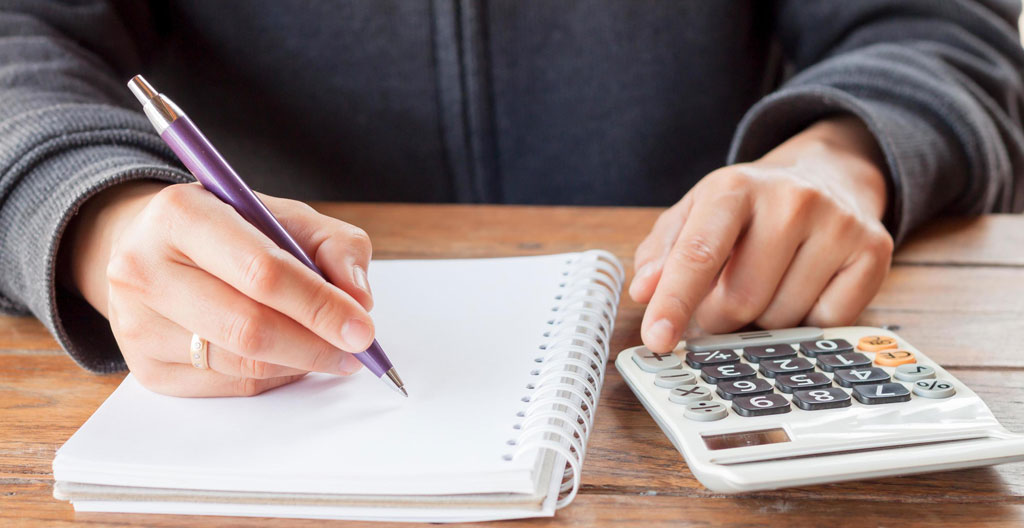 Gros plan sur une main tenant un stylo au-dessus d'un carnet avec des chiffres manuscrits, calculatrice visible en arrière-plan flouté