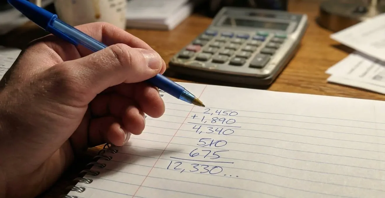 Gros plan sur une main tenant un stylo au-dessus d'un carnet avec des chiffres manuscrits, calculatrice visible en arrière-plan flouté