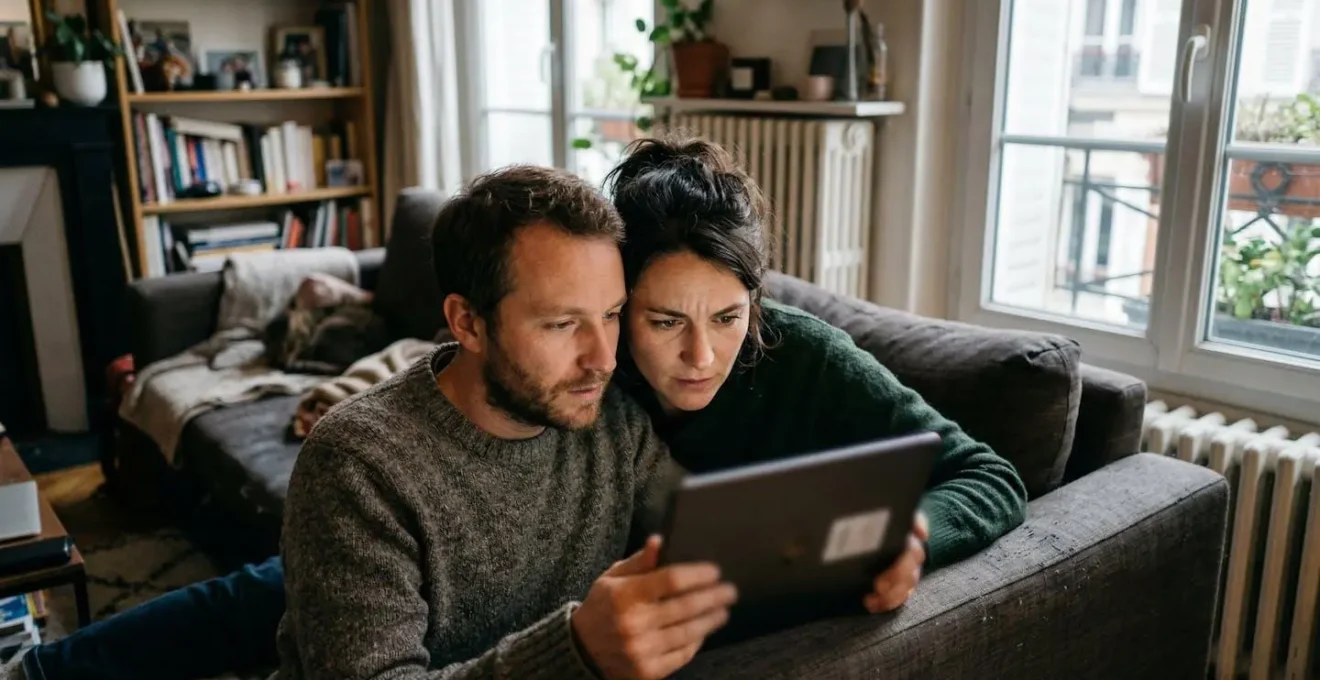 Un couple trentenaire consulte une tablette sur le canapé de leur salon moderne, expressions concentrées et détendues